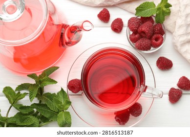 Refreshing raspberry tea in glass cup, teapot, fresh berries and mint leaves on white wooden table, flat lay