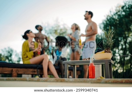 Refreshing red beverage sits in the foreground, slightly blurred, while in the background, a group of friends enjoys a poolside gathering, creating a vibrant summer scene