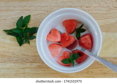 Refreshing slices of red, ripe and juicy heart-shaped watermelon in a white bowl with a fork and mint leaves on a light wooden background top view