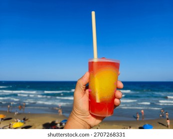 A refreshing summer drink held by a hand against a vibrant blue sky and ocean backdrop at the beach.