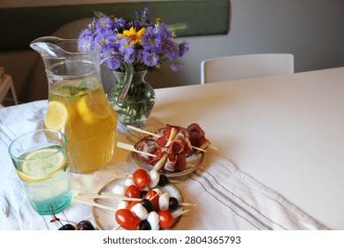 Refreshing summer table setting with a pitcher of lemonade, colorful skewers of appetizers, and a vase of flowers on a white tablecloth in a bright indoor space