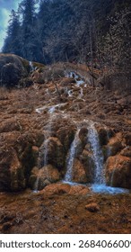 A refreshing valley view where spring water cascades gently over mossy rocks, surrounded by pine forested hills, creating a serene and invigorating morning scene.