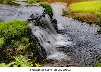 A refreshing view of a small waterfall cascading over a lush green, mossy rocky ledge into a peaceful river in a forest landscape during summer.