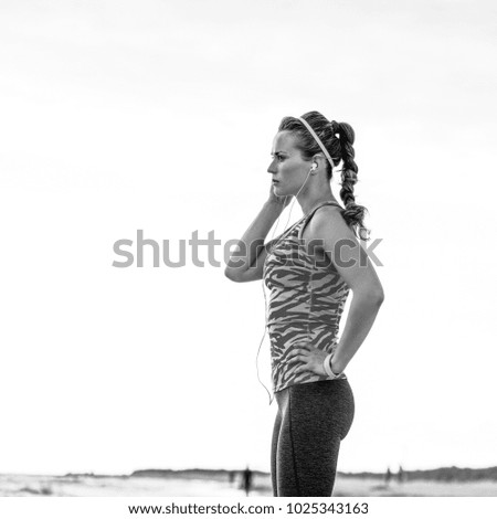 Refreshing wild sea side workout. Full length portrait of pensive fit woman in sport clothes on the seashore looking into the distance and listening to the music with headphones