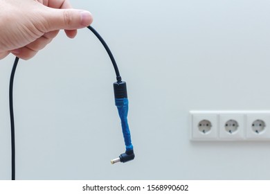 Refurbished Laptop Power Adapter Wire In Hand On Wall Background With Sockets. Danger Of Short Circuit And Electric Shock