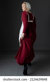 A Regency Maid Servant Wearing A Red Linen Dress With An Apron And A Cap Against A Studio Backdrop