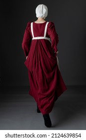 A Regency Maid Servant Wearing A Red Linen Dress With An Apron And A Cap Against A Studio Backdrop