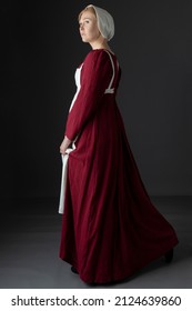 A Regency Maid Servant Wearing A Red Linen Dress With An Apron And A Cap Against A Studio Backdrop