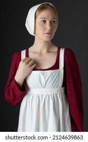 A Regency Maid Servant Wearing A Red Linen Dress With An Apron And A Cap Against A Studio Backdrop