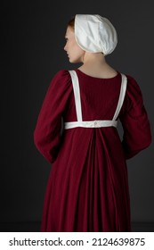 A Regency Maid Servant Wearing A Red Linen Dress With An Apron And A Cap Against A Studio Backdrop