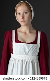 A Regency Maid Servant Wearing A Red Linen Dress With An Apron And A Cap Against A Studio Backdrop