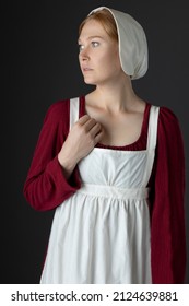 A Regency Maid Servant Wearing A Red Linen Dress With An Apron And A Cap Against A Studio Backdrop