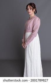 A Regency woman wearing a cream embroidered dress and a pink spencer posing against a grey backdrop