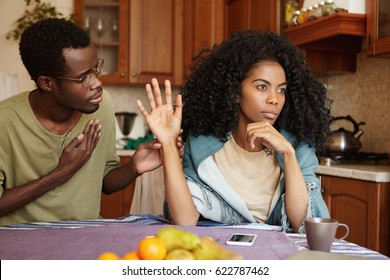 Regretful unhappy young African American male in glasses trying hard to sweet talk his mad offended wife who is sitting next to him at kitchen table, refusing all his lies. People and relationships - Powered by Shutterstock