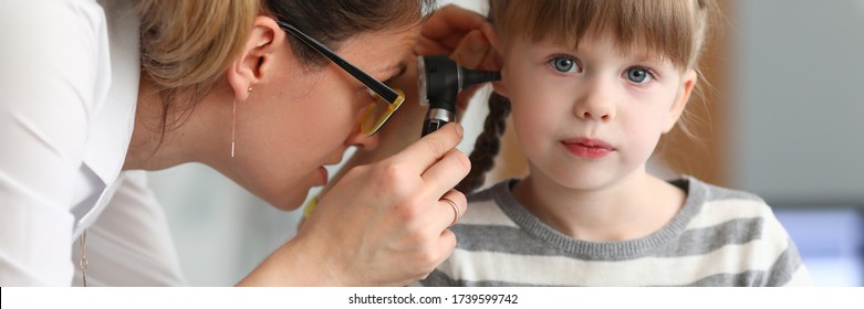 Regular pediatric medical examination at doctor. Female ENT wears glasses and jewelry, she examines ear small patient with special device. Girl sits quietly on couch and awaits end procedure.