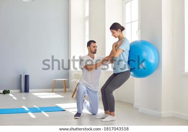 Rehabilitation\
after injury. Physiotherapist helping woman with backache to\
restore spinal health after physical trauma. Young female athlete\
doing back exercise using soft yoga\
ball