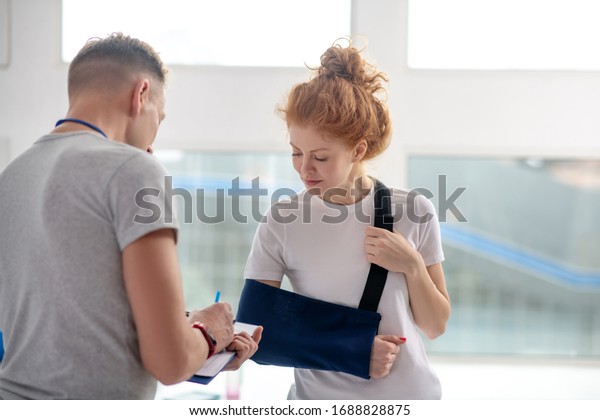 At\
rehabilitation center. Male physiotherapist writing instructions,\
female patient standing in front of him in arm\
sling