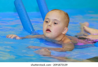 Rehabilitation Of A Child With Down Syndrome In A Pool, A Boy Swims In Blue Water.
