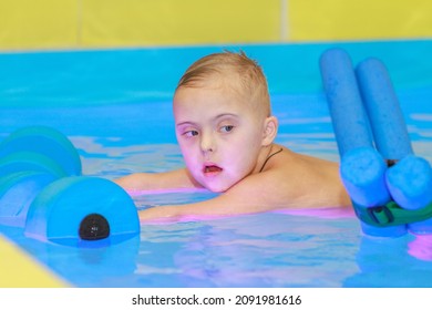 Rehabilitation Of A Child With Down Syndrome In A Pool, A Boy Swims In Blue Water.
