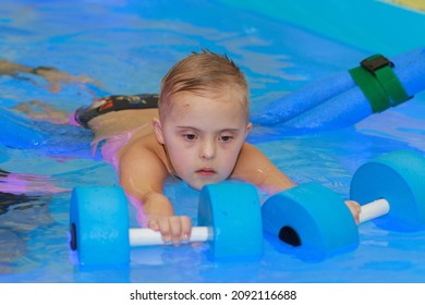 Rehabilitation Of A Child With Down Syndrome In A Pool, A Boy Swims In Blue Water.
