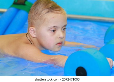 Rehabilitation Of A Child With Down Syndrome In A Pool, A Boy Swims In Blue Water.
