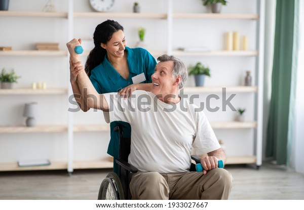 Rehabilitation of disabled\
people concept. Young physiotherapist helping senior male patient\
in wheelchair exercise at home. Handicapped elderly man training\
with dumbbells