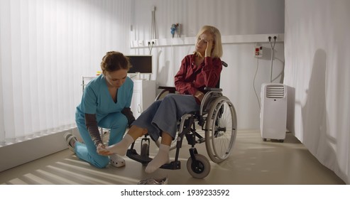 Rehabilitation Doctor Helping To Lift Leg Of Patient In Wheelchair. Nurse Massaging Feet Of Senior Paralyzed Woman Sitting In Wheelchair In Hospital Room