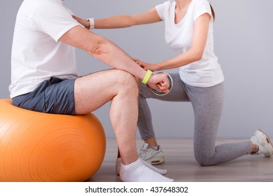 Rehabilitation Together. Cropped Image Of Male Patient Sitting On A Gym Ball With Dumbbells During Physical Therapy Praxis And Female Physiotherapist Exercising With Him