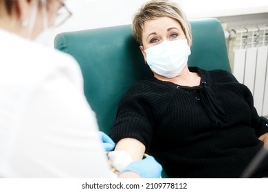 Rehabilitation And Treatment In Medical Clinic. 40-50 Year Old Woman In Medical Mask Sits In Chair And Looks Into Camera. Nurse Injects Woman Into Vein.