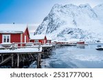 Reine village with traditional rorbu houses on Lofoten islands in winter. Scandinavian landscape with red wooden fishermen cabins on frozen sea water and snowy Reinebringen mountain, northern Norway.