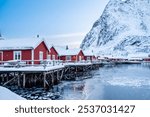 Reine village with traditional rorbu houses on Lofoten islands in winter. Scandinavian landscape with red wooden fishermen cabins on frozen sea water and snowy Reinebringen mountain, northern Norway.