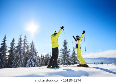 Rejoicing Young Woman And Man On Skis With Hands Up With Ski Poles On Wooded Snow-covered Mountain Top On Winter Sunny Day At Ski Resort. Activity, Thrill, Victory, Achievement Concept. Copy Space.