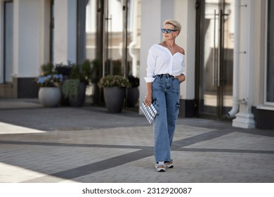 Rejuvenated Trendy mature woman with short hair stands outdoors in city street wears youth clothing white shirt, jeans cargo pants, clutch. Urban style fashion, age and timeless beauty.