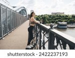 Rejuvenated young Caucasian woman taking a rest on a city bridge railing, smartphone in hand with earphones, after a revitalizing run, overlooking a serene riverscape