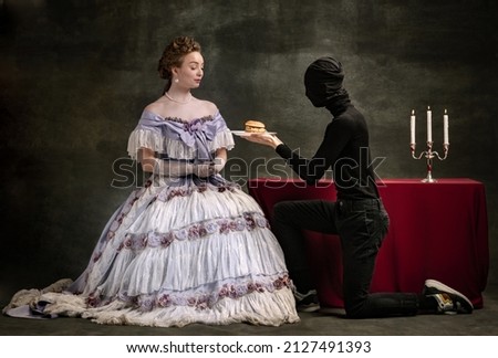 Relation. Cinematic portrait of young beautiful woman in image of medieval royal person in renaissance style dress and her servant, page isolated on dark vintage background. Comparison of eras