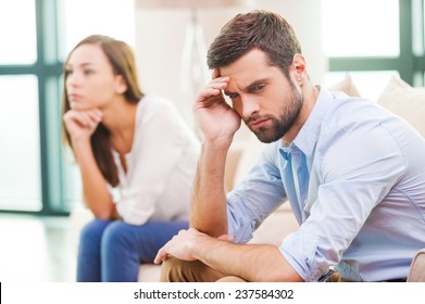 Relationship Breakdown. Depressed Young Man Holding Hand On Head And Looking Away While Woman Sitting Behind Him On The Couch 