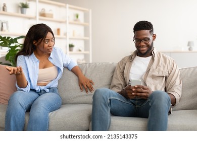 Relationship Conflicts Concept. Annoyed Angry Black Woman Looking And Yelling At Her Man Who Using Cellphone And Ignoring His Confused Lady, Couple Sitting On Couch In Living Room