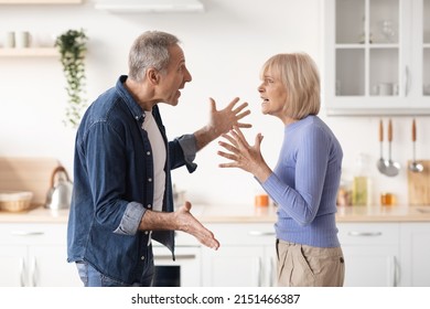 Relationship Problems Concept. Angry Senior Man Arguing With His Wife At Kitchen While Cooking Dinner, Husband And Wife Shouting At Each Other And Gesturing, Side View, Copy Space