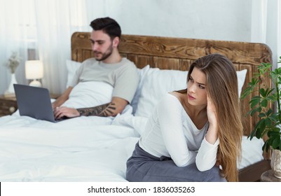 Relationship, problems, a lot of work and virtual treason. Upset woman sitting on bed and looking at husband, texting on laptop or working in modern rustic bedroom interior in evening, free space
