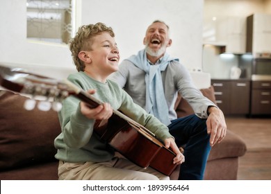 Relationships between the younger and older generations. Happy grandfather and his excited little grandson sitting together on sofa, laughing and enjoying playing guitar, spending time at home - Powered by Shutterstock