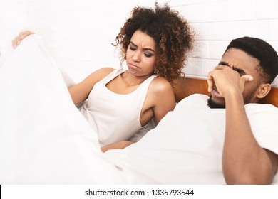 Relationships difficulties, stress and impotence. Black couple having marital problems. Woman in bed looking under blanket, man with erectile dysfunction