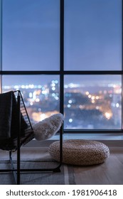 Relax Area In Front Of Big Panoramic Window With Night City View. Vertical Shot Of Modern Metal Armchair With Fur Hygge Throw And Wicker Ottoman In Living Room. Modern Furniture In Contemporary Design