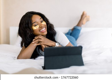 Relax and beauty procedures on weekend. Pleasant African smiling woman lying on bed in cozy room, with golden eye patches under her eyes and watching a film on ipad tablet or having a video call - Powered by Shutterstock