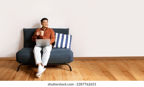 Relax concept. Happy asian man drinking coffee using pc sitting on comfortable sofa and dreaming, panorama with free space. Male resting on sofa, enjoying weekend free time - Powered by Shutterstock