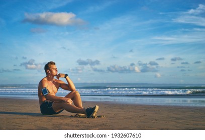 Relax And Enjoy Music. Young Handsome Man With Earphones And Smartphone Sitting On The Beach.