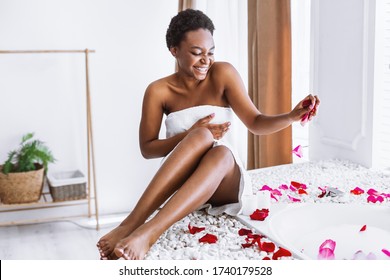 Relax in evening. Smiling african american girl in towel makes bath with foam and petals - Powered by Shutterstock