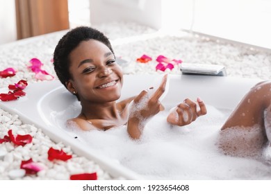 Relax, fun and rest in spare time at home during covid-19 lockdown. Cheerful millennial african american cute lady in bath with petals, plays with foam in bathroom interior in morning, copy space