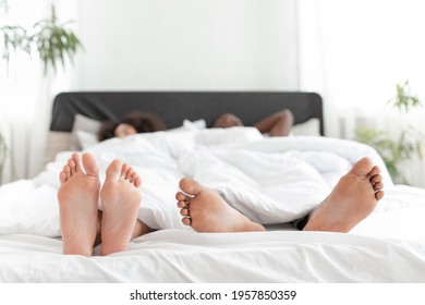 Relax On Weekend, Romance Together And Lazy Couple At Free Time In Morning. Happy Millennial African American Female And Guy Lie In Bed Under Blanket In Bedroom Interior, Feet Close Up, Empty Space