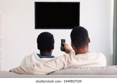 Relax. Rear Back View Of Black Couple Watching Tv, Man Holding And Using Remote Controller, Looking At Blank Television On The Wall With Isolated Empty Monitor For Mock Up Template, Selective Focus