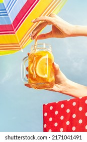 Relax Time. Female Hand Holding Glass Mug With Frosty Orange Lemonade, Cocktail Over Blue Sunny Sky Background. Vacation, Happiness, Summer Vibes. Retro Style Photo. Tropical Climate